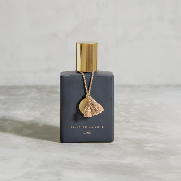 MATTE BLACK GLASS BOTTLE WITH SHINY GOLD CAP. GOLD METAL DISC HANGING FROM BOTTLE NECK ON A TAUPE SILK STRING. WRITING ON BOTTLE IN GOLD READS FILLE DE LA LUNE MANON