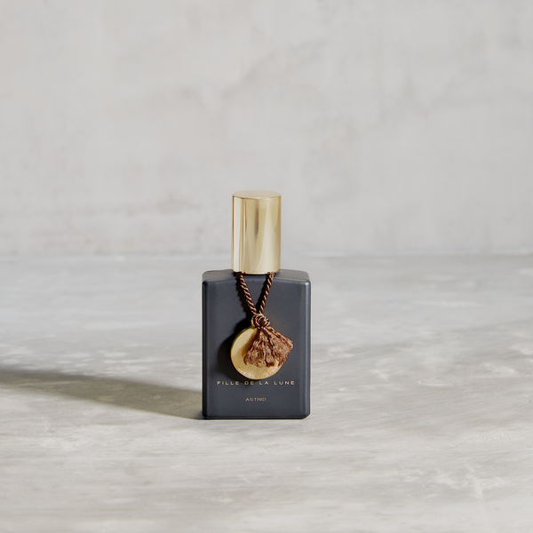 MATTE BLACK GLASS BOTTLE WITH SHINY GOLD CAP. GOLD METAL DISC HANGING FROM BOTTLE NECK ON A TAUPE SILK STRING. WRITING ON BOTTLE IN GOLD READS FILLE DE LA LUNE  ASTRID