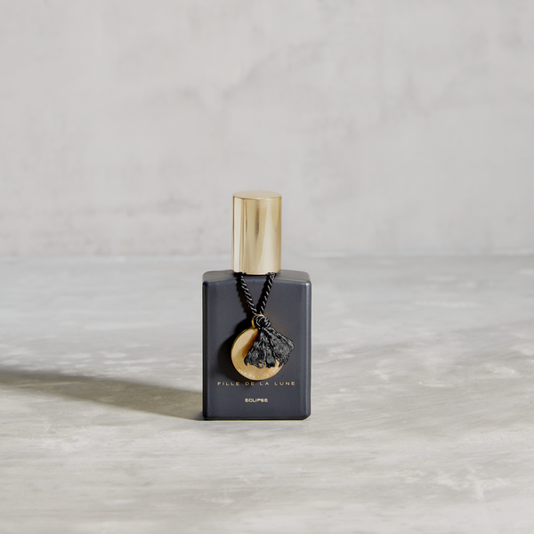 MATTE BLACK GLASS BOTTLE WITH SHINY GOLD CAP. GOLD METAL DISC HANGING FROM BOTTLE NECK ON A BLACK SILK STRING. WRITING ON BOTTLE IN GOLD READS FILLE DE LA LUNE ECLIPSE