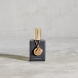 MATTE BLACK GLASS BOTTLE WITH SHINY GOLD CAP. GOLD METAL DISC HANGING FROM BOTTLE NECK ON A TAUPE SILK STRING. WRITING ON BOTTLE IN GOLD READS FILLE DE LA LUNE SOLSTICE D'ETE