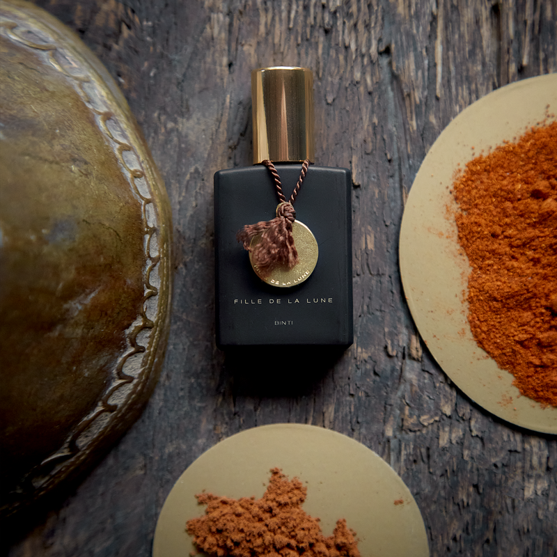 MATTE BLACK GLASS BOTTLE WITH SHINY GOLD CAP. GOLD METAL DISC HANGING FROM BOTTLE NECK ON A TAUPE SILK STRING. WRITING ON BOTTLE IN GOLD READS FILLE DE LA LUNE BINTI. BOTTLE IS ON TOP OF AGED WOODEN TABLE . TO THE RIGHT OF IT IS A PLATE OF ORANGE SPICES. BELOW IT IS A SMALLER PLATE OF ORANGE SPICES. TO THE LEFT OF THE PERFUME BOTTLE IS AN ANTIQUE GOLD BELL.