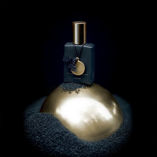 MATTE BLACK GLASS BOTTLE WITH SHINY GOLD CAP. GOLD METAL DISC HANGING FROM BOTTLE NECK ON A BLACK SILK STRING. WRITING ON BOTTLE IN GOLD READS FILLE DE LA LUNE ECLIPSE. THE BOTTLE IS ON TOP OF A GOLD METAL BALL THAT IS SURROUNDED BY TINY BLACK PEBBLES. THE BACKGROUND IS A DARK BLACK SHADOW.