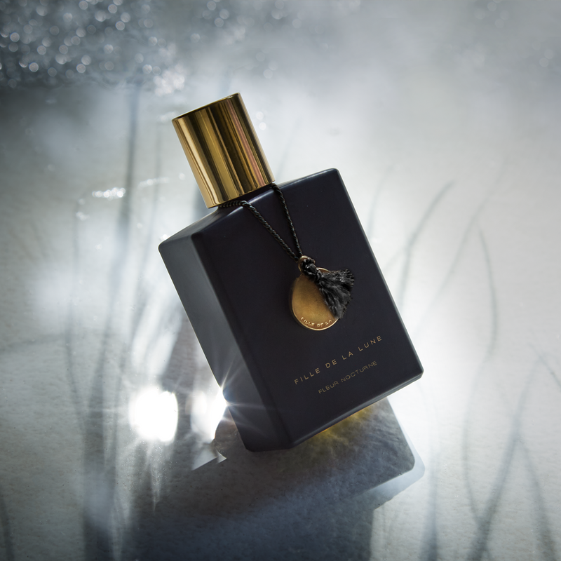 MATTE BLACK GLASS BOTTLE WITH SHINY GOLD CAP. GOLD METAL DISC HANGING FROM BOTTLE NECK ON A BLACK SILK STRING. WRITING ON BOTTLE IN GOLD READS FILLE DE LA LUNE FLEUR NOCTURNE. THE BOTTLE IS SURROUNDED BY AND ON A SILVER REFLECTIVE TABLE