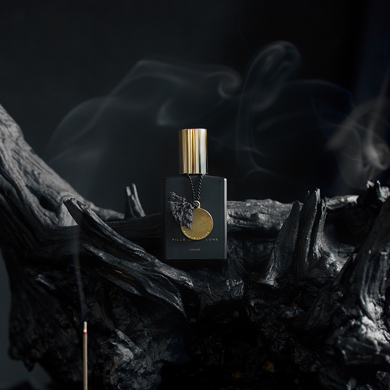 MATTE BLACK GLASS BOTTLE WITH SHINY GOLD CAP. GOLD METAL DISC HANGING FROM BOTTLE NECK ON A BLACK SILK STRING. WRITING ON BOTTLE IN GOLD READS FILLE DE LA LUNE SACRE. BOTTLE IS LAYING AGAINST DARK AGED WOOD. IN FRONT IS A SINGLE BURNING INCENSE THAT IS LETTING OUT SMOKE WHICH SURROUNDS THE ATMOSPHERE.