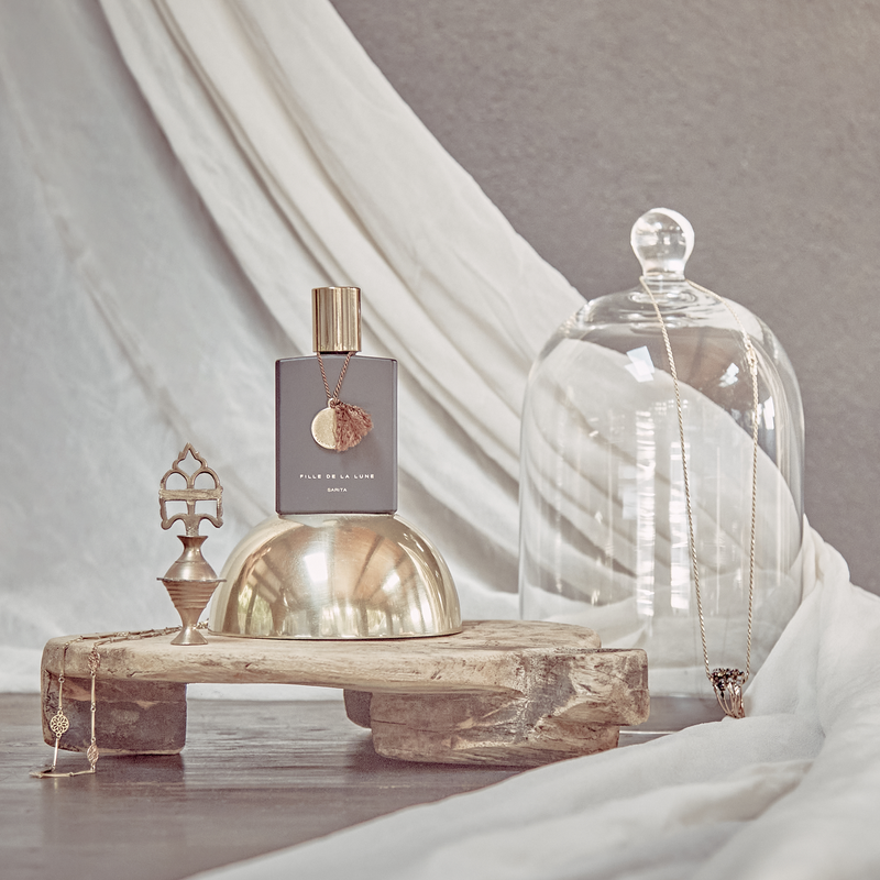 MATTE BLACK GLASS BOTTLE WITH SHINY GOLD CAP. GOLD METAL DISC HANGING FROM BOTTLE NECK ON A TAUPE SILK STRING. WRITING ON BOTTLE IN GOLD READS FILLE DE LA LUNE SARITA. BOTTLE IS ON TOP OF A  GOLD METAL HALF SPHERE. UNDERNEATH IT IS A SMALL WOODEN STAND. TO THE LEFT OF THE BOTTLE IS AN ANTIQUE FIGURE. TO THE RIGHT OF THE BOTTLE IS A CLEAR GLASS COVER WITH JEWELRY HANGING FROM IT.