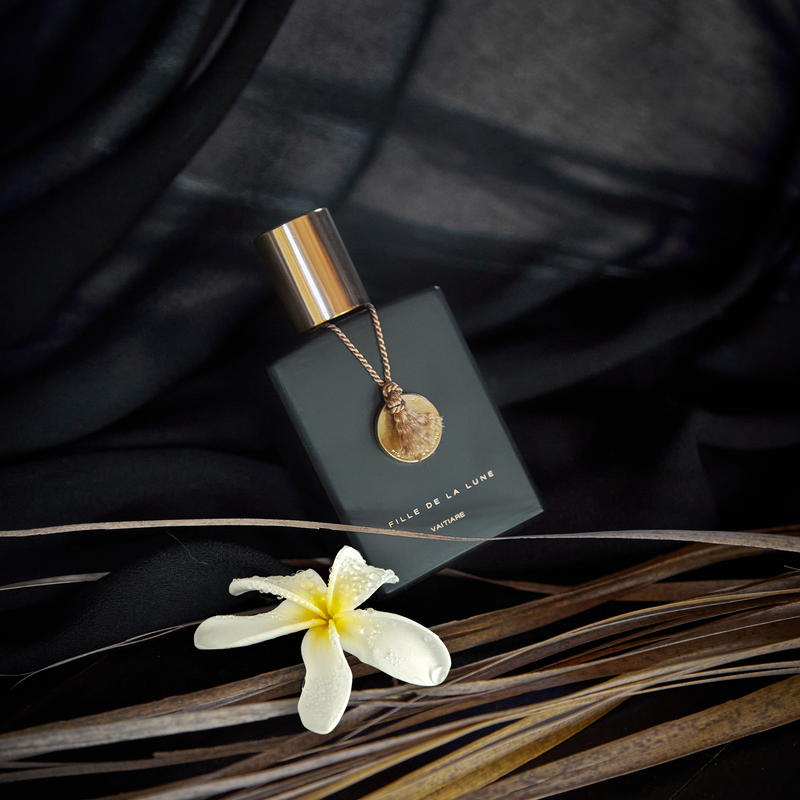 MATTE BLACK GLASS BOTTLE WITH SHINY GOLD CAP. GOLD METAL DISC HANGING FROM BOTTLE NECK ON A TAUPE SILK STRING. WRITING ON BOTTLE IN GOLD READS FILLE DE LA LUNE VAITIARE. THE BOTTLE IS LAYING ON BLACK FABRIC. IN FRONT OF IT IS ONE WHITE FLOWER WHICH SITS ON TOP OF DRIED BROWN LEAVES.