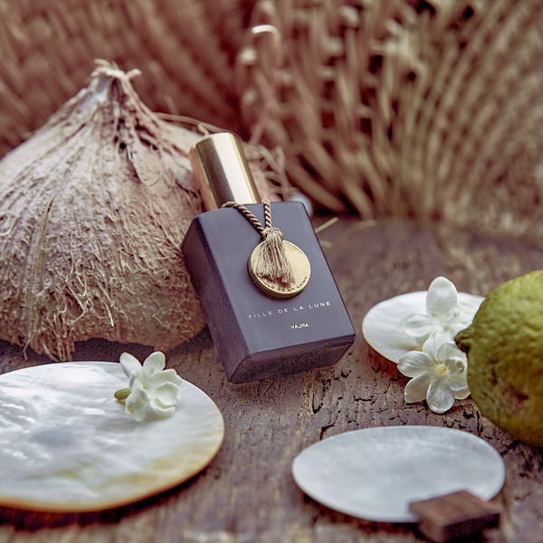 MATTE BLACK GLASS BOTTLE WITH SHINY GOLD CAP. GOLD METAL DISC HANGING FROM BOTTLE NECK ON A TAUPE SILK STRING. WRITING ON BOTTLE IN GOLD READS FILLE DE LA LUNE (BRAND NAME) VAJRA (PERFUME NAME). BOTTLE LAYING AGAINST SHELL OF COCONUT ON TOP OF AGED WOODEN TABLE WITH THREE PEARL DISCS AND WHITE FLOWERS SURROUNDING BOTTLE. PART OF GREEN LIME ON TABLE. BACKDROP OF STRAW FANS.
